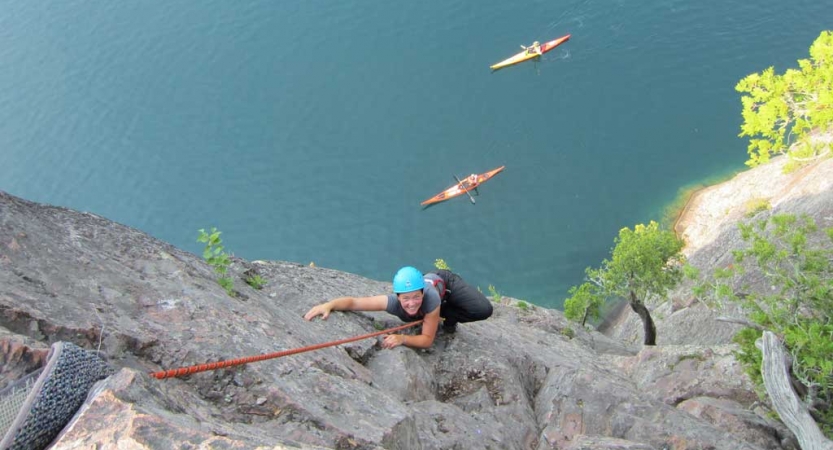 A person wearing safety gear and secured by ropes pauses their climb to look up at the camera. Below them, two kayaks rest on vast blue water. 
